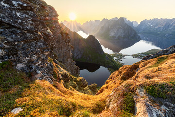 The midnight sun pierces the sky above Reinebringen, illuminating the moss-draped rocks and casting a radiant glow over the dramatic peaks and fjords of Lofoten Island