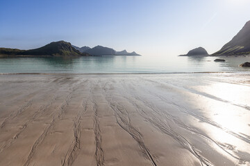 Tidal patterns etch the sands of Haukland Beach, Lofoten, Norway, leading to tranquil waters under the glow of a bright summer sun