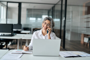 Happy middle aged business woman talking on phone using laptop in office. Smiling mature older female bank manager, senior professional lady making business call on telephone working sitting at desk. - Powered by Adobe
