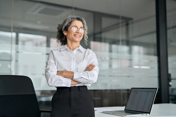 Happy older professional business woman standing in office looking away. Smiling middle aged mature...