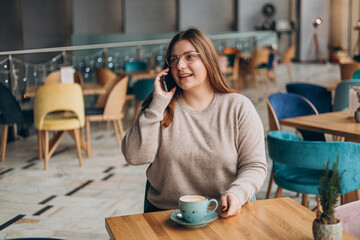 Attractive 30s female with cup of coffee having talking conversation with mobile phone while rest in cafe. Casual young woman using mobile phone indoors