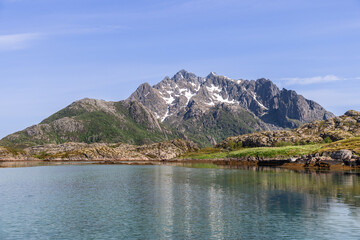 A serene vista of Lofoten, Norway, where rugged peaks adorned with snow patches tower above a tranquil, reflective fjord under a clear blue sky