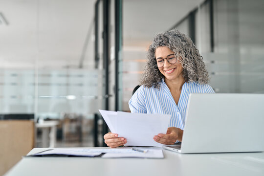 Busy smiling mature older business woman working in office checking document. Middle aged female hr consultancy manager, notary or lawyer using laptop reading paper cv law financial file, tax invoice.