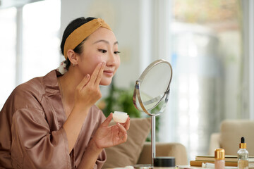 Smiling young woman looking at magnifying mirror when applying hydrating face cream