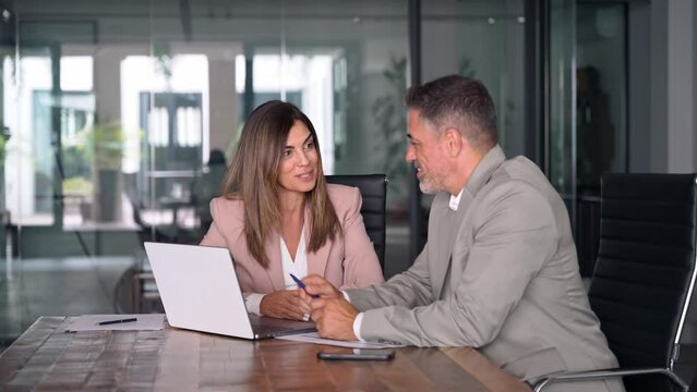Smiling middle aged professional business woman executive working looking at laptop computer discussing digital data with male colleague at office meeting planning corporate strategy.