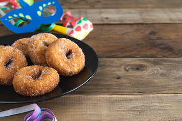 Sweet doughnuts on a black plate on a wooden background with typical carnival decorations. Copy...