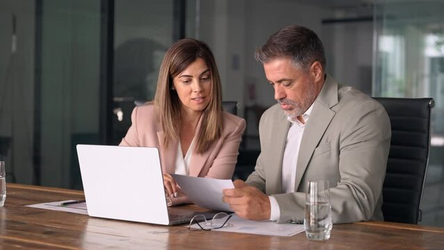 Two happy professional business partners executive leaders managers wearing suits working in office looking at laptop computer discussing documents together at corporate meeting.