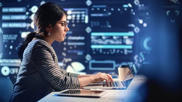 Indian woman programmer working overtime in dark office of digital company, typing text on laptop
