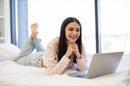 Happy smiling brunette female watching movie using modern laptop, staying at home. Charming woman in casual attire enjoying reading information on Internet while lying on cozy bed.