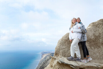 Pretty women stand on a high mountain on the sea coast on a sunny day. Smiling blondes hugging. Active lifestyle and recreation. Full height.
