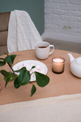 Home kitchen interior - white teapot with hot tea, cup of tea, plate with marshmellow, burning candle, green plant in ceramic pot, napkin, beige chair on green background.