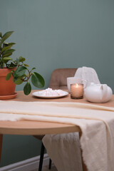 Home kitchen interior - white teapot with hot tea, cup of tea, plate with marshmellow, burning candle, green plant in ceramic pot, napkin, beige chair on green background.