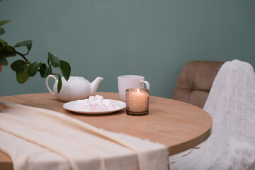 Home kitchen interior - white teapot with hot tea, cup of tea, plate with marshmellow, burning candle, green plant in ceramic pot, napkin, beige chair on green background.