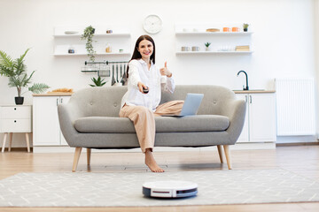 Young caucasian brunette woman with laptop programming automatic tidying of room floor. Beautiful lady switching on robot vacuum cleaner via remote control while sitting on sofa in kitchen.