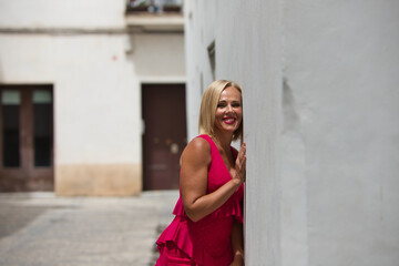 Beautiful blonde mature woman in an elegant red dress leaning against the white wall of a building on a narrow street in cadiz, andalusia, spain. The woman is happy and smiling.