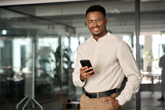 Smiling busy professional African American business man standing in office holding mobile cellphone. Young Black businessman entrepreneur using smartphone looking at camera with cell phone at work.