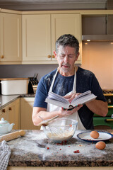 Man looks at a cookery book in the kitchen. Wearing an apron, surrounded by ingredients and mess. Learning how to bake is a new challenge for a mature man. Real life skills concept. Cookery school. 