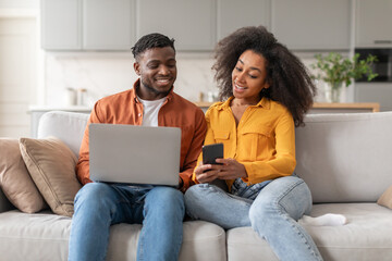 Happy Black Couple Reading Message On Smartphone Using Laptop Indoors
