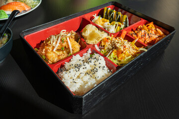 Japanese Lunch in Bento Box with Deep-Fried Squid Rings, Sushi Rolls, Salad, Rice, Pickled Ginger