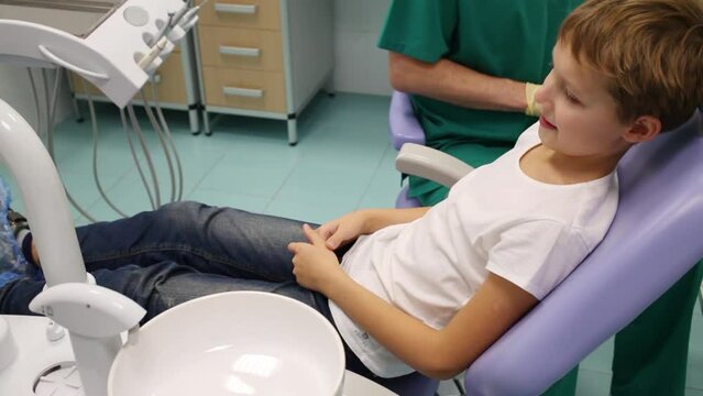 Little boy is sitting on armchair near dental drill and doctor at hospital.