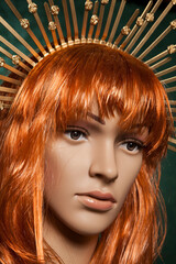 Mannequin with bright long red hair wearing a spiky golden crown posing on a green background