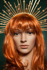Mannequin with bright long red hair wearing a spiky golden crown posing on a green background