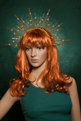 Mannequin with bright long red hair wearing a spiky golden crown posing on a green background