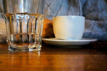 Weiße Tasse Kaffee neben Glas Wasser auf brauner Holztischplatte vor sitzender junger Frau mit weißem Pullover aus Wolle in Café am Nachmittag im Winter
