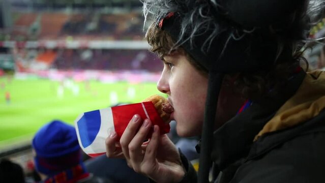 Teenager in a fur hat eating a hot dog at the stadium 