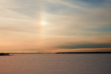 A winter rainbow (halo) over a frozen snow-covered river. In the distance a factory with many smoking pipes. Copy space.