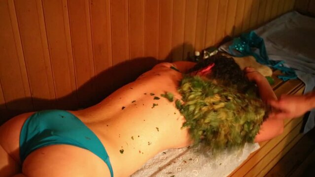 Woman lies on the bench in the bath, person smacks her with a broom