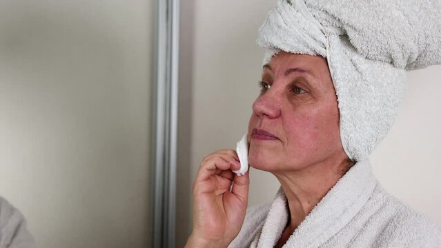 beautiful elder old woman granny is applying face cream,use cotton disk and hydrofil oil to remove makeup.female grandmother looking in double sided mirror against wall, dressed in bath robe 4k