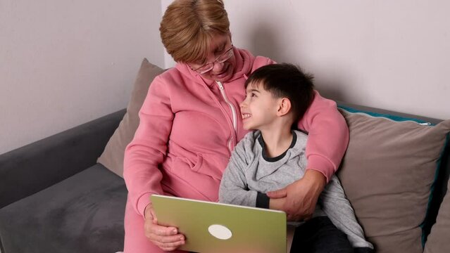 beautiful older granny and nephew sitting on sofa in front of laptop,talking to someone. grandmother old female lady with vision glasses on eyes and smiling kid child boy. togetherness concept 4k