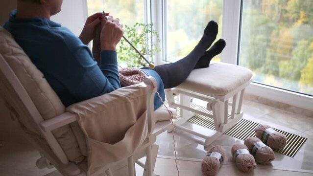 Woman sits in a chair with ottoman and knitting, yarn lying on the floor