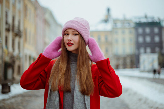 Fashionable happy smiling woman wearing pink beanie hat, mittens, red coat, gray scarf, posing in street of European city. Christmas, outdoor winter holidays concept. Copy, empty, blank space for text