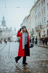 Fashionable confident woman wearing elegant red midi coat, gray cap, scarf, turtleneck, black leather gloves, wide leg jeans, walking in street of European city. Outdoor full-length fashion portrait