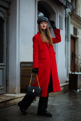 Fashionable confident woman wearing elegant red midi long coat, gray cap, black gloves, jeans,...