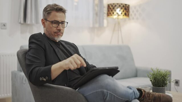 Business portrait - Businessman at home working with tablet computer. Home office, browsing internet. Mature age, middle age, mid adult man in 50s sitting in armchair in living room.