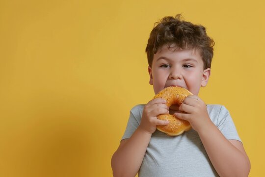 Cute little boy eating a hamburger on a yellow background. Child with obesity. Overweight and obesity concept. Obesity Concept with Copy Space.