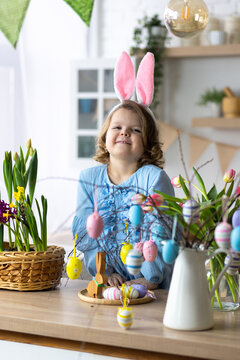Portrait of cute pretty smiling little girl in blue dress and bunny rabbit ears preparing easter table decor. Fresh spring bright colourful flowers in the kitchen. Sunny day, family time