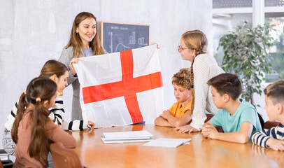 Young female teacher showing flag of England to schoolchildren preteens during history lesson in...