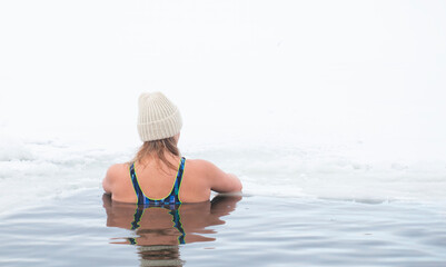 A woman in a swimsuit and a warm hat swims in an ice hole during winter swimming, winter therapy,...