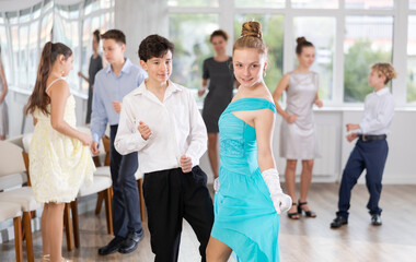 Smiling teenage girl in elegant dress having fun at school prom with group of peers, dancing with...