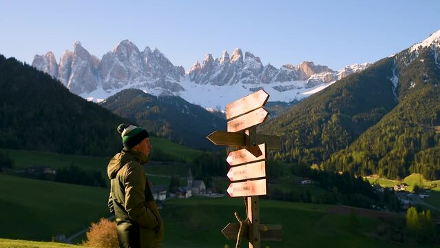 Man tourist looking at wooden pointer at the trail in Dolomites Alps near Santa Maddalena village. Male traveler looking for path to medieval church in Val Di Funes, Dolomiti, Alto Adige, Italy