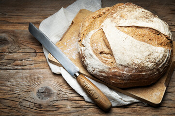 Whole loaf of traditional bread with knife, cutting board and tea towel on wooden background, close-up, copy space.