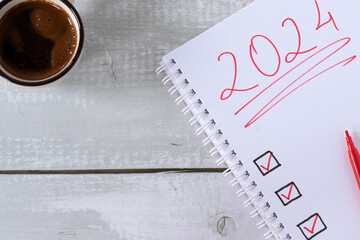 2024 handwritten number in a notebook with a checklist and coffee cup on wood. Top table view. New...