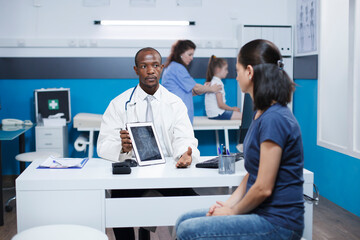 Seated in hospital office, African American male physician with a tablet is having a consultation with a female patient. Reassuring atmosphere, equipped with medical equipment.