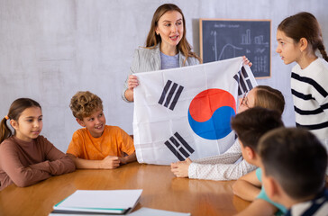 Joyful young female teacher showing flag of South Korea to schoolchildren preteens during history lesson in classroom