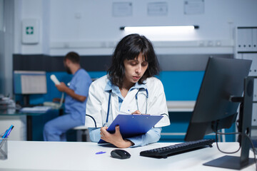 Female doctor preparing for medical consultations using a clipboard and computer. Image shows...