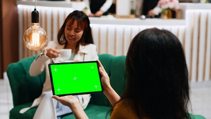 Traveler checking isolated greenscreen template on tablet, sitting with her friend in lounge area and looking at blank copyspace template. Asian guest using isolated mockup. Tripod shot.